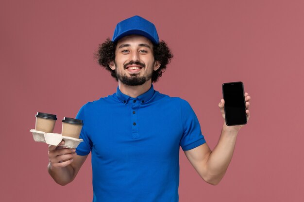 Vorderansicht des männlichen Kuriers in blauer Uniform und Kappe mit Kaffeetassen und Arbeitstelefon auf seinen Händen an der rosa Wand