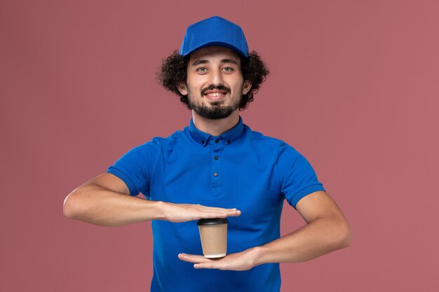 Vorderansicht des männlichen Kuriers in blauer Uniform und Kappe mit Lieferkaffeetasse auf seinen Händen auf rosa Wand