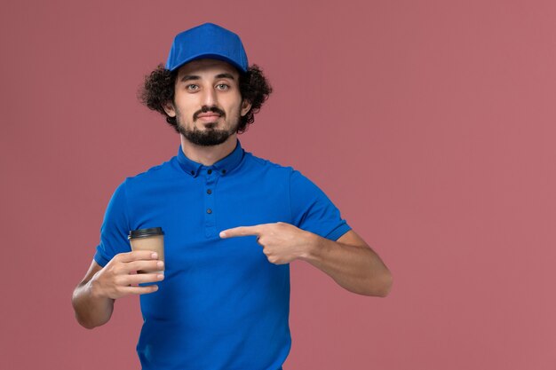 Vorderansicht des männlichen Kuriers in blauer Uniform und Kappe mit Lieferkaffeetasse auf seinen Händen auf rosa Wand