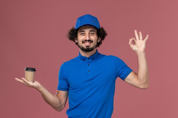 Vorderansicht des männlichen Kuriers in blauer Uniform und Kappe mit Lieferkaffeetasse auf seinen Händen auf rosa Wand