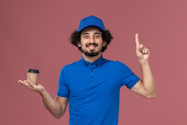 Vorderansicht des männlichen Kuriers in blauer Uniform und Kappe mit Lieferkaffeetasse auf seinen Händen auf rosa Wand