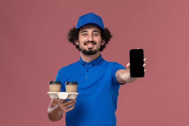 Vorderansicht des männlichen Kuriers in blauer Uniform und Kappe mit Telefon- und Lieferkaffeetassen auf seinen Händen auf rosa Wand