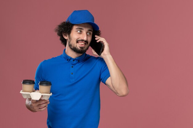 Vorderansicht des männlichen Kuriers in der blauen Uniform und in der Kappe mit den Kaffeetassen der Lieferung auf seinen Händen, die am Telefon auf rosa Wand sprechen
