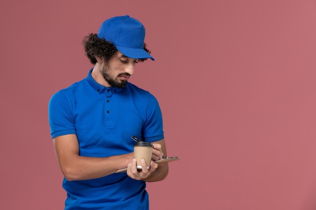 Vorderansicht des männlichen Kuriers in der blauen Uniform und in der Kappe mit der Kaffeetasse und dem Notizblock auf seinen Händen, die Notizen auf der rosa Wand schreiben