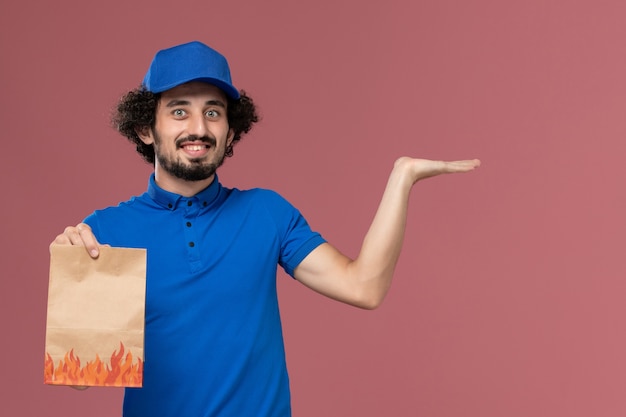 Vorderansicht des männlichen Kuriers in der blauen Uniformkappe mit dem Lebensmittelpaket der Lieferpapier auf seinen Händen, die auf der hellrosa Wand aufwerfen