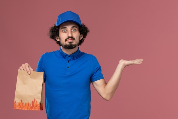 Vorderansicht des männlichen Kuriers in der blauen Uniformkappe mit dem Lebensmittelpaket des Lieferpapiers auf seinen Händen auf der hellrosa Wand