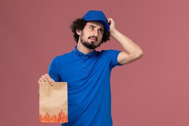 Vorderansicht des männlichen Kuriers in der blauen Uniformkappe mit dem Lebensmittelpaket des Lieferpapiers auf seinen Händen auf der hellrosa Wand