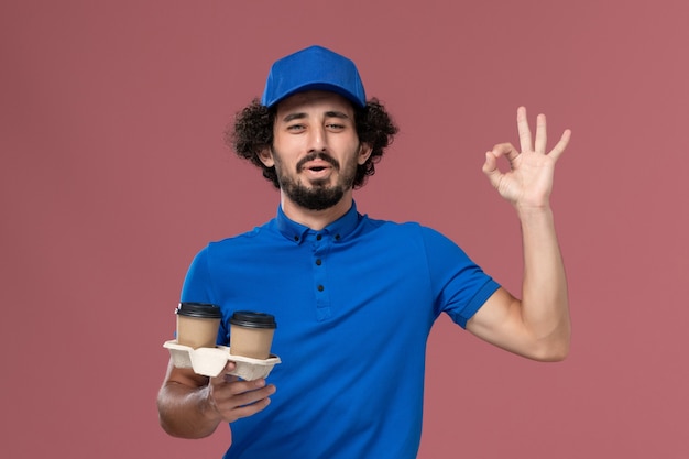 Vorderansicht des männlichen Kuriers in der blauen Uniformkappe mit den Kaffeetassen der Lieferung auf seinen Händen auf der rosa Wand