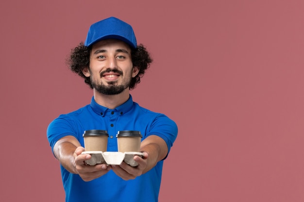 Vorderansicht des männlichen Kuriers in der blauen Uniformkappe mit den Kaffeetassen der Lieferung auf seinen Händen auf hellrosa Wand