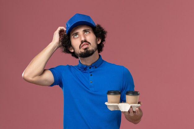Vorderansicht des männlichen Kuriers in der blauen Uniformkappe mit den Kaffeetassen der Lieferung auf seinen Händen und dem Denken an der rosa Wand