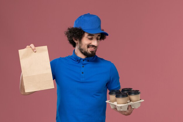 Vorderansicht des männlichen Kuriers in der blauen Uniformkappe mit den Kaffeetassen und der Lebensmittelverpackung auf seinen Händen auf der rosa Wand