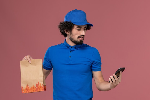 Vorderansicht des männlichen Kuriers in der blauen Uniformkappe mit Liefernahrungsmittelpaket auf seinen Händen unter Verwendung des Telefons an der rosa Wand