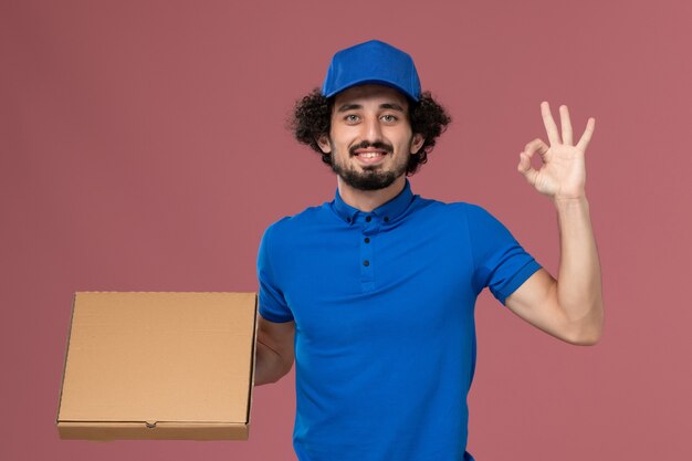 Vorderansicht des männlichen Kuriers in der blauen Uniformkappe mit Nahrungsmittelbox auf seinen Händen auf hellrosa Wand