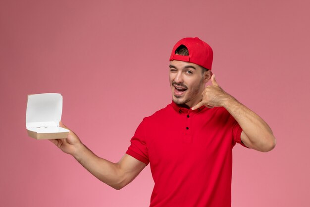 Vorderansicht des männlichen Kuriers in der roten Uniform und in der Kappe, die kleines Lieferpaket an der rosa Wand halten