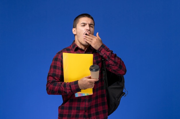 Vorderansicht des männlichen Schülers im roten karierten Hemd mit dem Rucksack, der gelbe Akten und den gähnenden Kaffee an der blauen Wand hält