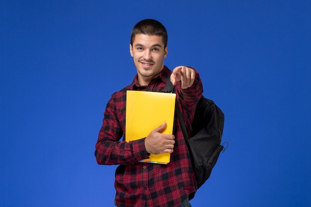 Vorderansicht des männlichen Schülers im roten karierten Hemd mit dem Rucksack, der gelbe Dateien an der blauen Wand hält