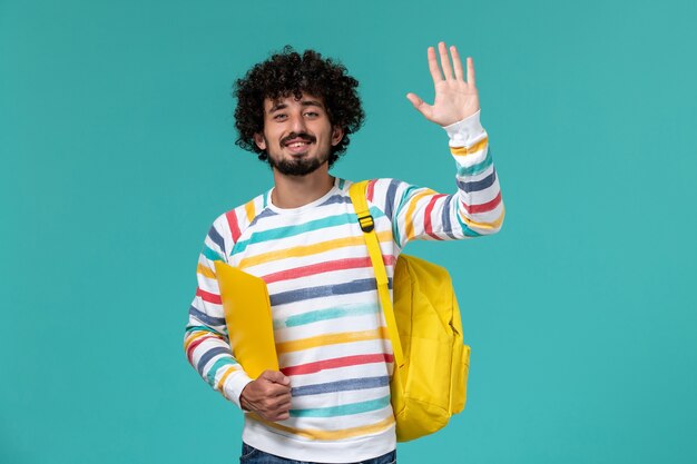 Vorderansicht des männlichen Studenten, der gelben Rucksack hält, der Dateien auf blauer Wand hält