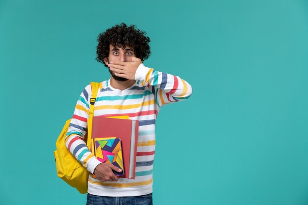 Vorderansicht des männlichen Studenten, der gelben Rucksack trägt, der Dateien und Hefte an der blauen Wand hält