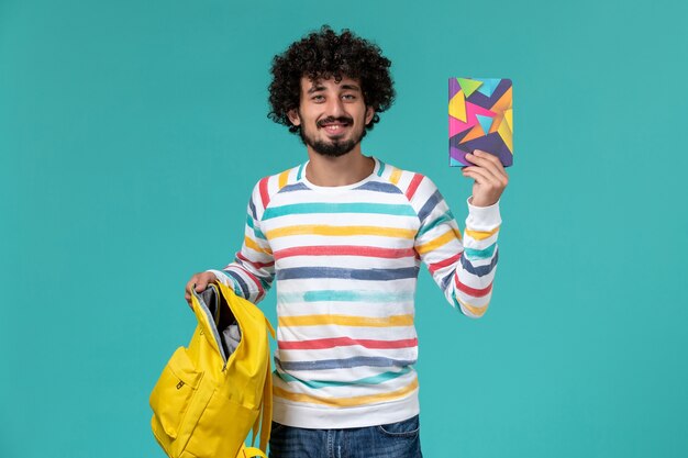 Vorderansicht des männlichen Studenten, der gelben Rucksack und Heft an der blauen Wand hält