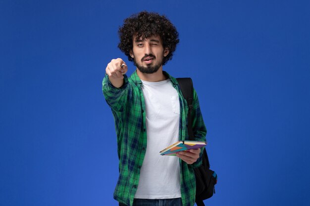 Vorderansicht des männlichen Studenten, der schwarzen Rucksack hält, der Heft und Stift an der blauen Wand hält