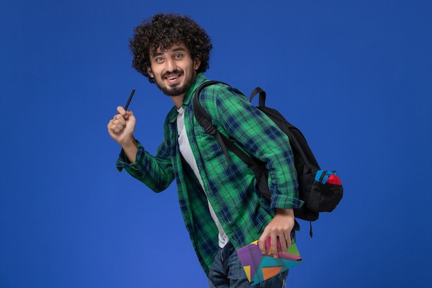 Vorderansicht des männlichen Studenten, der schwarzen Rucksack hält, der Heft und Stift hält, der auf blaue Wand läuft