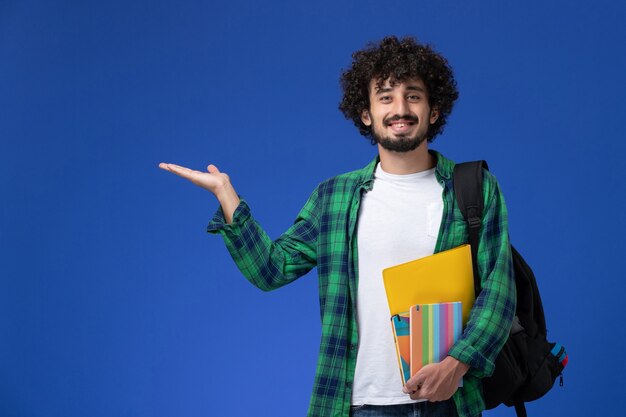 Vorderansicht des männlichen Studenten, der schwarzen Rucksack trägt, der Hefte hält und an der blauen Wand lächelt