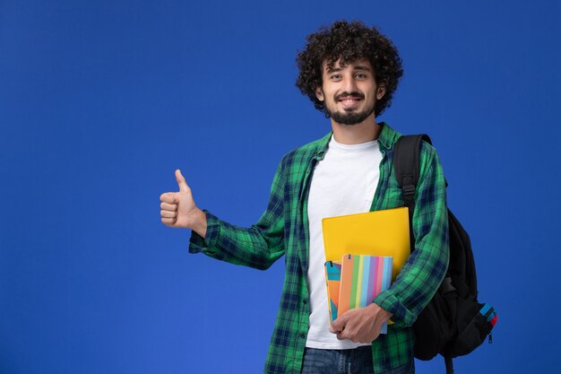 Vorderansicht des männlichen Studenten, der schwarzen Rucksack trägt, der Hefte hält und an der blauen Wand lächelt