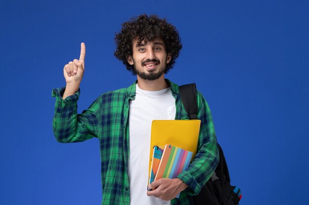 Vorderansicht des männlichen Studenten, der schwarzen Rucksack trägt, der Hefte und Akten an der blauen Wand hält