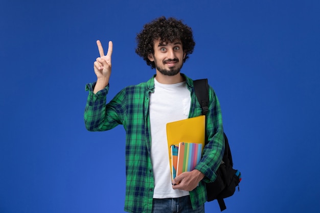 Vorderansicht des männlichen Studenten, der schwarzen Rucksack trägt, der Hefte und Akten an der blauen Wand hält