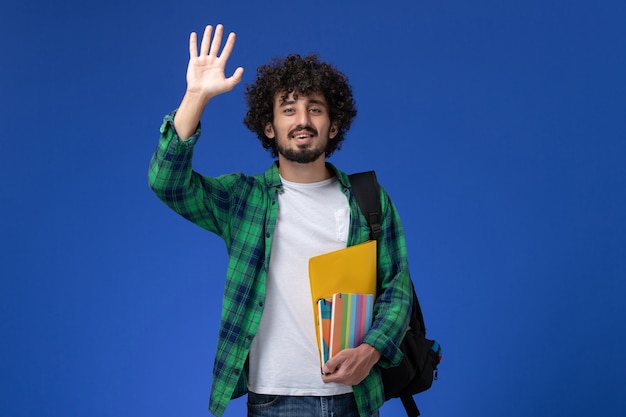 Vorderansicht des männlichen Studenten, der schwarzen Rucksack trägt, der Hefte und Akten hält, die seine Hand auf der blauen Wand winken