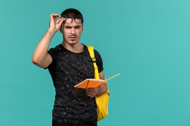 Vorderansicht des männlichen Studenten im gelben Rucksack des dunklen T-Shirts, der das Heft auf der blauen Wand hält und liest