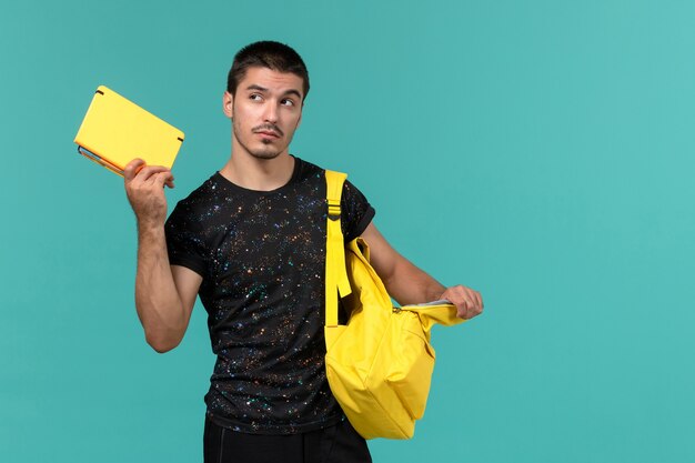 Vorderansicht des männlichen Studenten im gelben Rucksack des dunklen T-Shirts, der das Heft auf hellblauer Wand hält