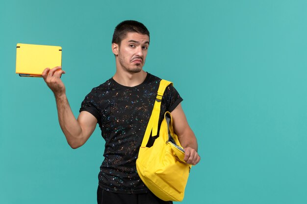 Vorderansicht des männlichen Studenten im gelben Rucksack des dunklen T-Shirts, der das Heft auf hellblauer Wand hält