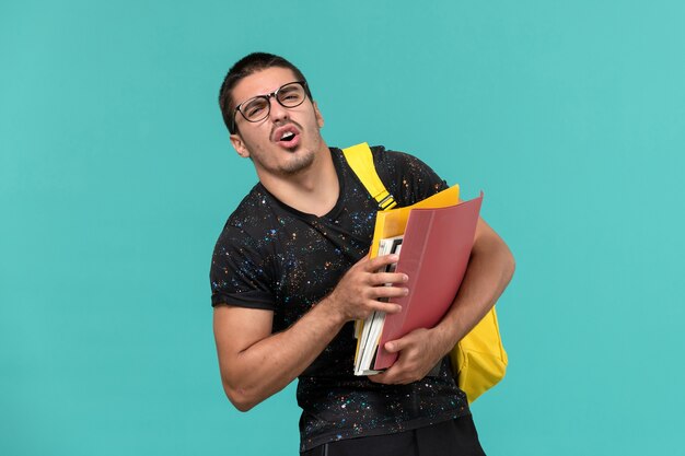 Vorderansicht des männlichen Studenten im gelben Rucksack des dunklen T-Shirts, der Dateien und Bücher auf der hellblauen Wand hält