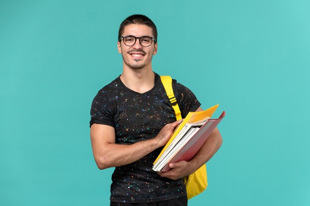 Vorderansicht des männlichen Studenten im gelben Rucksack des dunklen T-Shirts, der Dateien und Bücher auf der hellblauen Wand hält