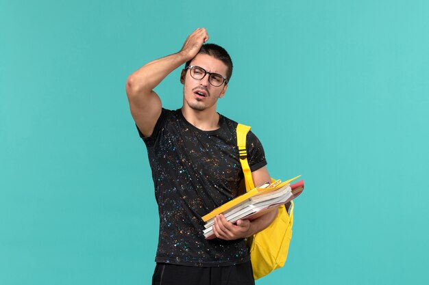 Vorderansicht des männlichen Studenten im gelben Rucksack des dunklen T-Shirts, der Dateien und Bücher hält, die an blaue Wand denken