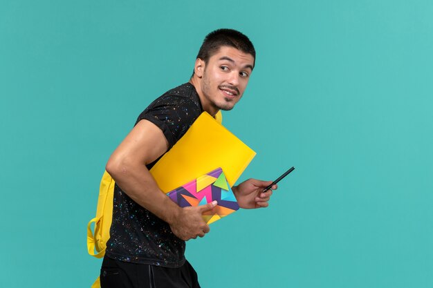 Vorderansicht des männlichen Studenten im gelben Rucksack des dunklen T-Shirts, der Dateien und Heft auf hellblauer Wand hält