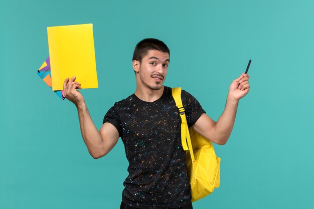 Vorderansicht des männlichen Studenten im gelben Rucksack des dunklen T-Shirts, der Dateien und Heft auf hellblauer Wand hält