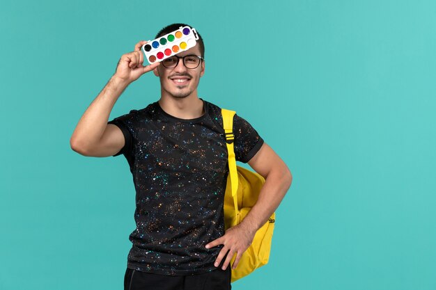 Vorderansicht des männlichen Studenten im gelben Rucksack des dunklen T-Shirts, der Farben hält, die auf blaue Wand lächeln