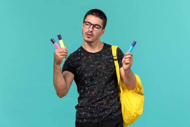 Vorderansicht des männlichen Studenten im gelben Rucksack des dunklen T-Shirts, der farbige Filzstifte auf blauer Wand hält