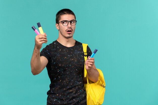 Vorderansicht des männlichen Studenten im gelben Rucksack des dunklen T-Shirts, der farbige Filzstifte auf blauer Wand hält