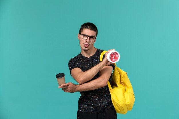 Vorderansicht des männlichen Studenten im gelben Rucksack des dunklen T-Shirts, der Kaffee und Uhren an der blauen Wand hält