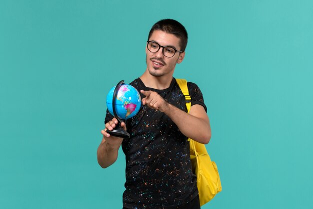 Vorderansicht des männlichen Studenten im gelben Rucksack des dunklen T-Shirts, der kleinen Globus auf der blauen Wand hält