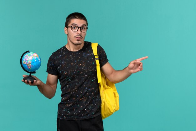 Vorderansicht des männlichen Studenten im gelben Rucksack des dunklen T-Shirts, der kleinen Globus auf der blauen Wand hält