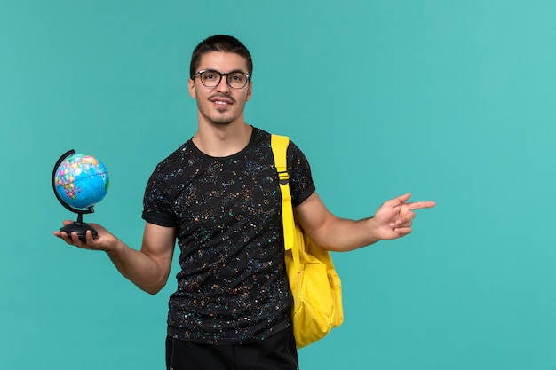 Vorderansicht des männlichen Studenten im gelben Rucksack des dunklen T-Shirts, der kleinen Globus hält, der auf blaue Wand lächelt