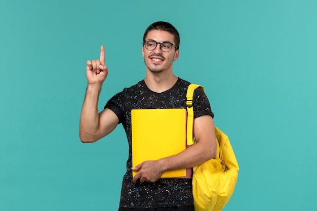 Vorderansicht des männlichen Studenten im gelben Rucksack des dunklen T-Shirts, der verschiedene Dateien auf der hellblauen Wand hält