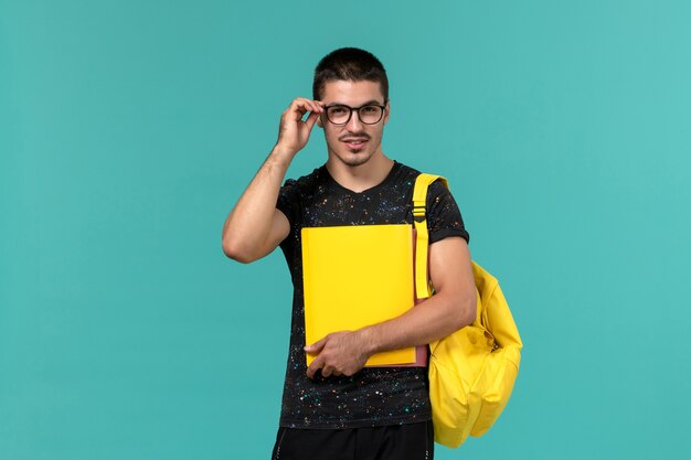 Vorderansicht des männlichen Studenten im gelben Rucksack des dunklen T-Shirts, der verschiedene Dateien auf der hellblauen Wand hält