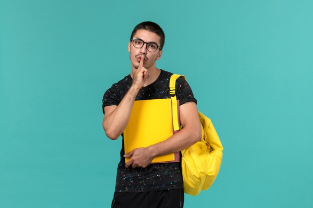 Vorderansicht des männlichen Studenten im gelben Rucksack des dunklen T-Shirts, der verschiedene Dateien auf der hellblauen Wand hält