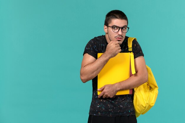 Vorderansicht des männlichen Studenten im gelben Rucksack des dunklen T-Shirts, der verschiedene Dateien auf der hellblauen Wand hält
