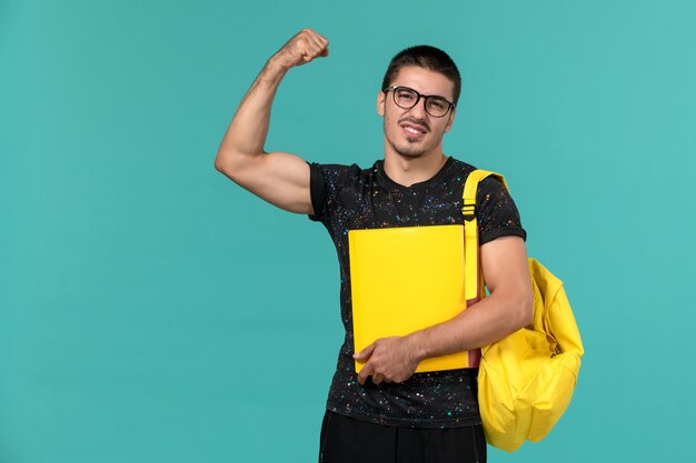 Vorderansicht des männlichen Studenten im gelben Rucksack des dunklen T-Shirts, der verschiedene Dateien hält, die an der hellblauen Wand biegen